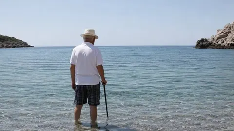Stariji muškarac u bijeloj košulji i šeširu stoji u plićaku aneg mora