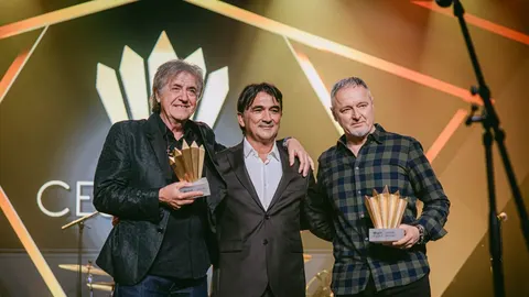 Trojica muškaraca u odjeći za svečanosti drže zlatne trofeje na pozornici
