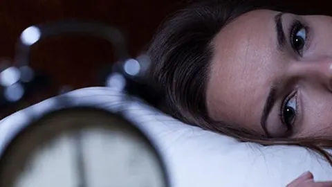 Žena s otvorenim očima leži na bijelom jastuku pored budilnika