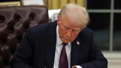 Donald Trump u tamnom odelu sedi u kožnoj fotelji i potpisuje dokument