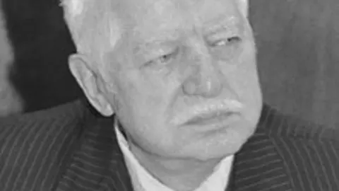 Crno-bijeli krupni plan starijeg muškarca ozbiljnog izraza lica u prugastom odijelu.