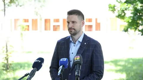Muškarac u plavom sako s mikrofonima ispred sebe na otvorenom