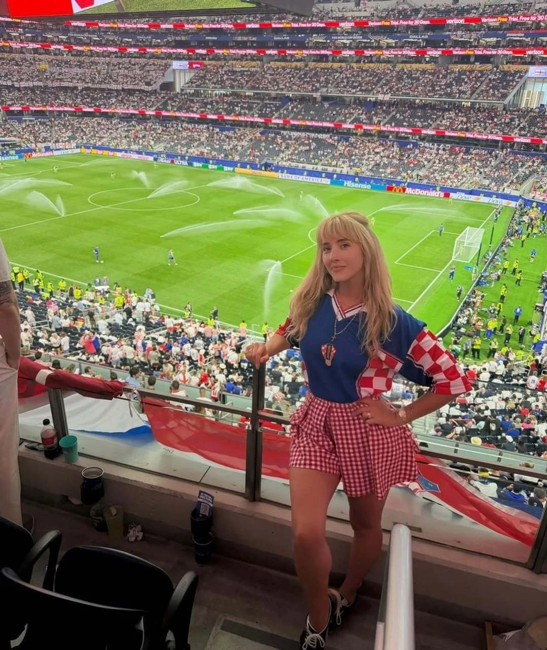 Plavokosa žena u crveno-bijelom dresu i suknji na nogometnom stadionu
