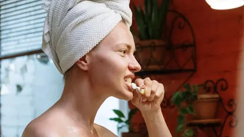 Žena s ručnikom na glavi pere zube četkicom u kupaonici