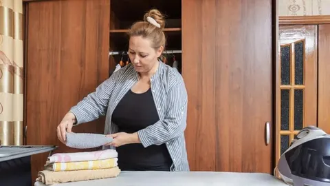 Žena slaže složene ručnike na dasku za peglanje u spavaćoj sobi