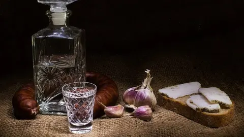 Kristalna čaša i bočica prozirne rakije uz kobasicu, češnjak i kruh sa slaninom