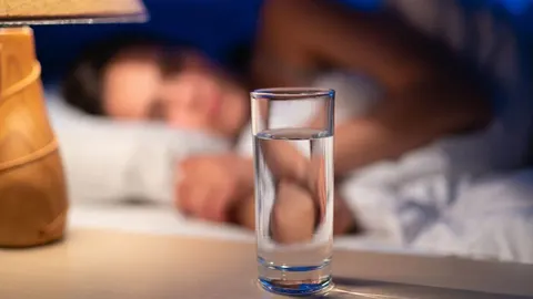 Čaša vode na noćnom stolicu s osobom koja spava u pozadini