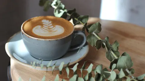 Šalica kave s pjenom i latte artom na drvenom stolu uz grančicu eukaliptusa