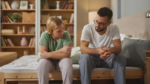 Mladi muškarac i žena sjede na rubu kreveta i razgovaraju u spavaćoj sobi.