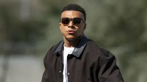 Kylian Mbappé u smeđoj jakni i sunčanim naočalama hoda na otvorenom.