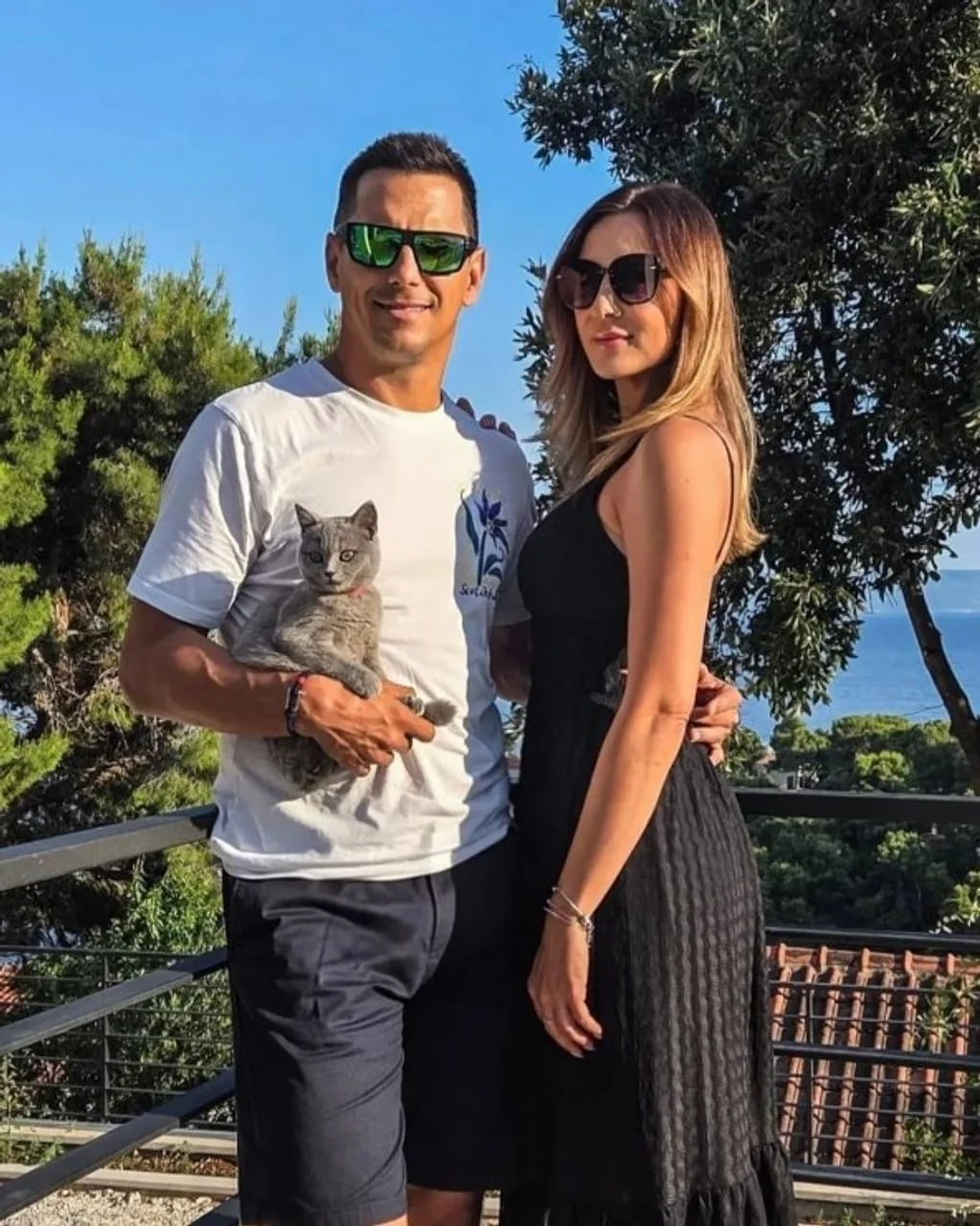Muškarac u bijeloj majici koji drži sivu mačku i žena u crnoj haljini stoje na terasi.