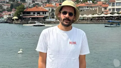 Muškarac u bijeloj majici i šeširu stoji ispred obale i grada