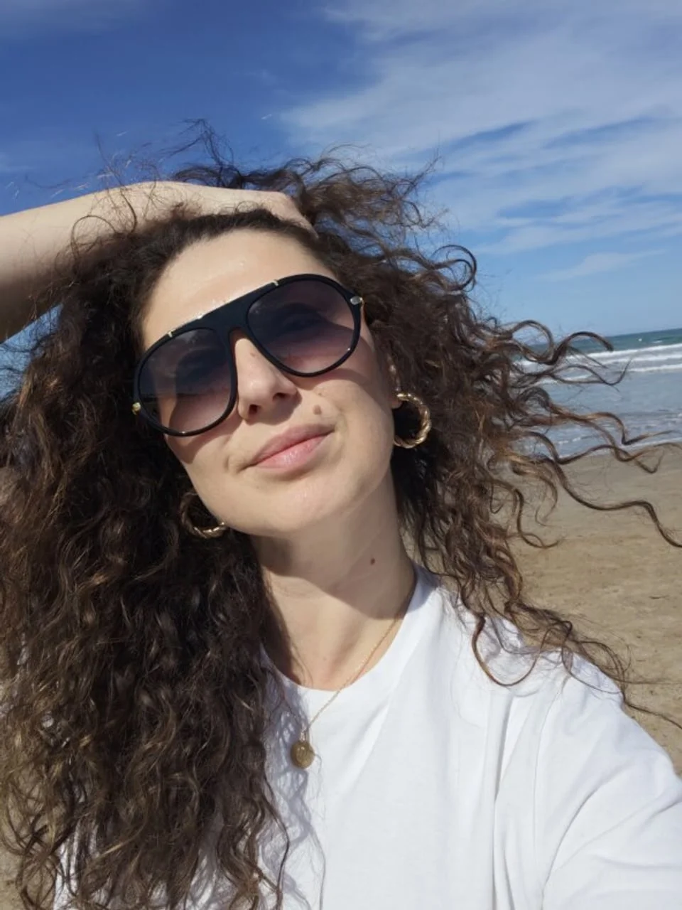 Portret žene s kovrčastom kosom, sunčalima i bijelom majicom na plaži