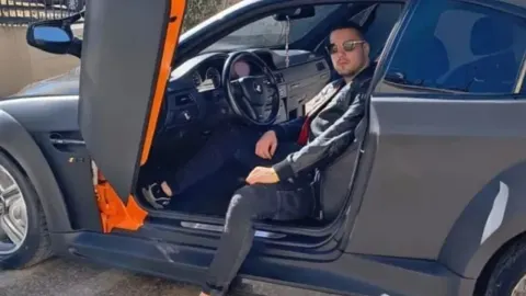 Mlađi muškarac sa sunčanim naočalama sjedi u sportskom automobilu s otvorenim vratima.