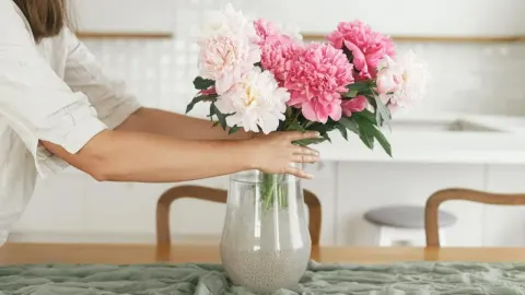 Žena stavlja buket ružičastih i bijelih božura u staklenu vazu na stolu