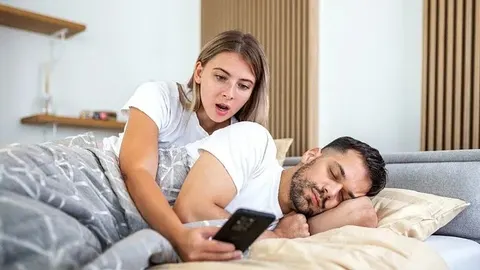 Žena iznenačeno gleda u mobitel dok muškarac spava pored nje u krevetu