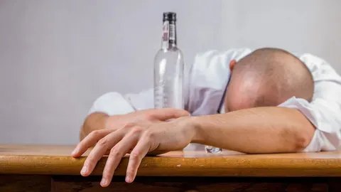 Muškarac u bijeloj košulji spava na drvenom stolu pored boce alkohola