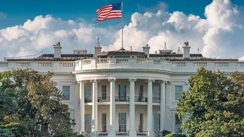 Bijela kuća u Washingtonu s američkom zastavom na vrhu pod plavim nebom.