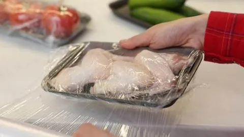 Ruka koja prekriva posudicu s sirovim pilećim krilcima prozirnom plastičnom folijom