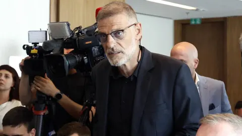 Slaven Bilić u tamnom odelu i naočalama okružen novinarima