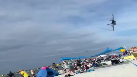 Borbeni avion u niskom letu nad pješčanom plažom s turistima