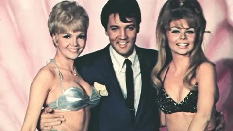 Elvis Presley u odelu između dve mlade žene u bikinijima
