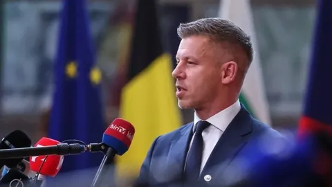 Portret Petera Magyara u tamnom odelu pred mikrofonima i zastavama