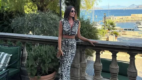 Žena u crno-bijelom uzorku stoji uz kamenu ogradu pored mora
