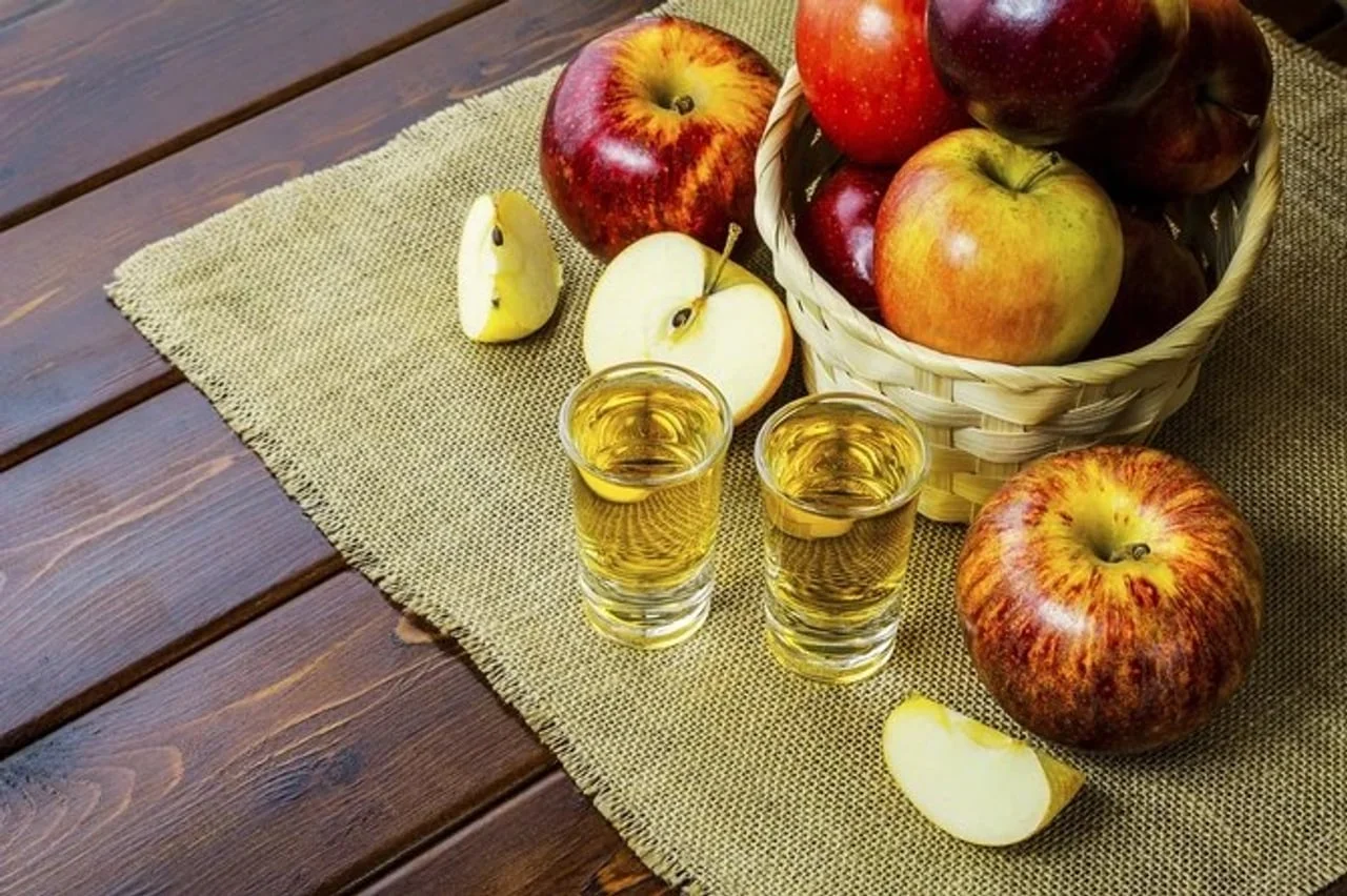 Dvije čašice žute tekućine pored košare punog crvenih jabuka na tkanini