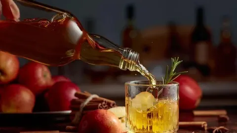 Sipanje žute tekućine iz staklene boce u čašu s jabukama u pozadini
