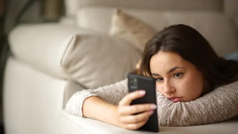 Tužna mlada žena leži na kauču i gleda u ekran pametnog telefona