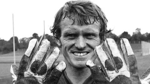 Nasmiješeni Sepp Maier pokazuje svoje vratarske rukavice na nogometnom terenu.