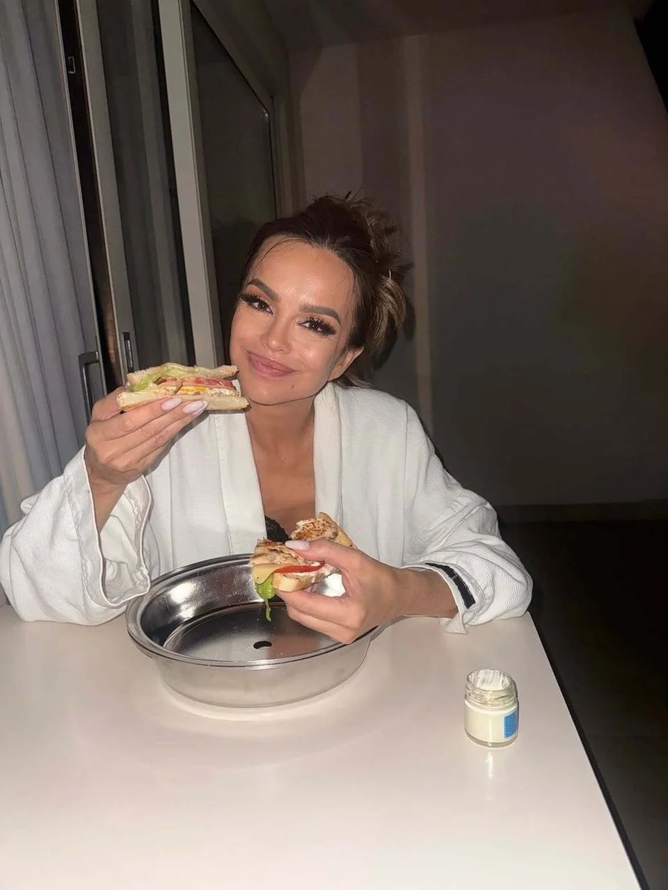 Severina u bijelom kućnom ogrtaču sjedi za stolom i drži sendvič