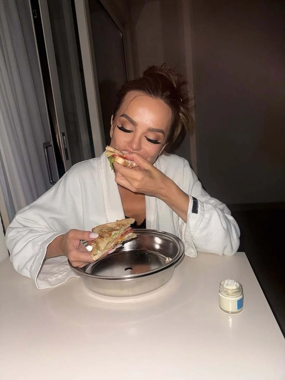 Severina u bijelom ogrtaču dok jede sendvič za bijelim stolom