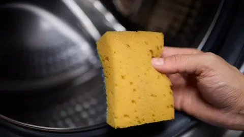 Ruka koja drži žutu kuhinjsku spužvu ispred otvorene sušilice rublja
