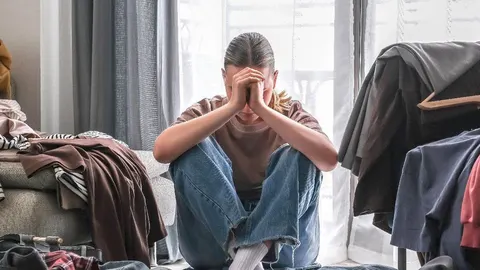 Žena sjedi na podu okružena hrpama neposloženog odjeće s rukama na licu