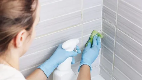 Žena u plavim rukavicama čisti pločice u kupaonici sprejem i spužvom