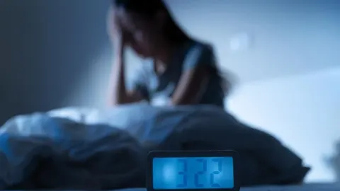 Žena sjedi na krevetu u mraku pored digitalne budilice koja pokazuje tri sata ujutro.