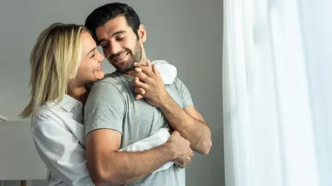 Nasmijani muškarac i žena u nježnom zagrljaju pored prozora.