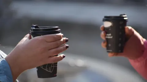 Krupni plan ruku koje drže crne papirnate šalice za kavu s crtežima.