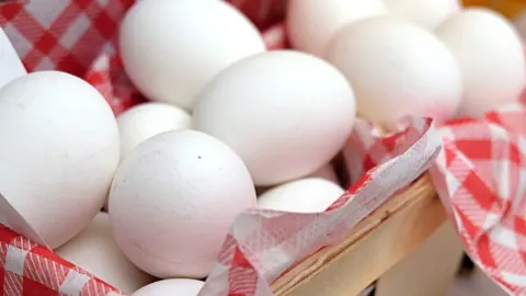 Krupni plan bijelih kokošjih jaja u drvenoj košari s crvenom kariranom krpom.