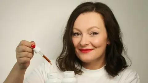 Žena drži pipetu s tamnom tekućinom iznad bijelih bočica suplemenata.