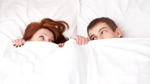 Muškarac i žena vire ispod bijelog pokrivača u spavaćoj sobi.