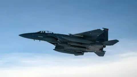 Moderni vojni lovački avion u letu iznad oblaka pod plavim nebom.