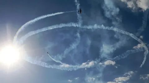 Zrakoplovi ostavljaju kružne bijele tragove na plavom nebu uz jarko sunce.