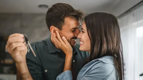 Nasmijani mladi par u zagrljaju dok muškarac pokazuje ključeve stana.