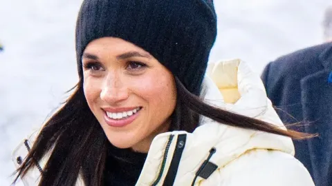 Meghan Markle nasmijana u bijeloj zimskoj jakni i crnoj kapi na snijegu.