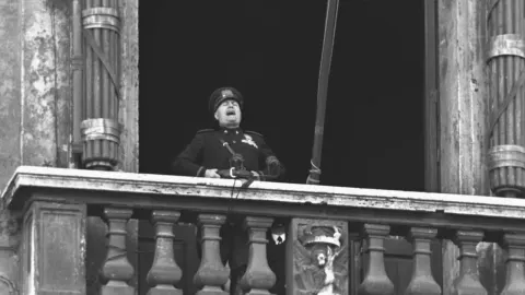 Crno-bijela fotografija Benita Mussolinija dok drži govor s balkona u uniformi.