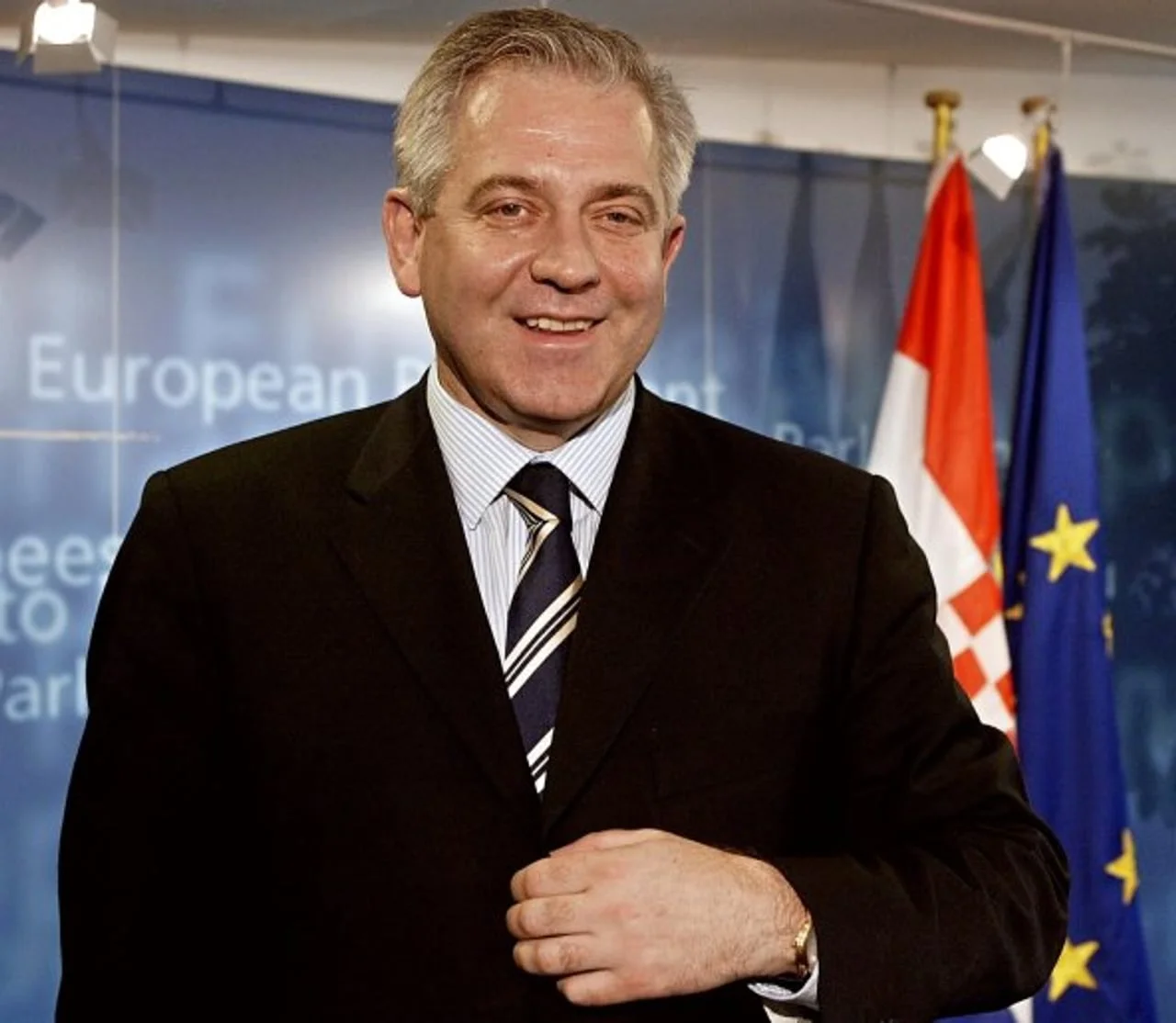 Nasmijani muškarac u odijelu ispred plave pozadine s europskim motivima.