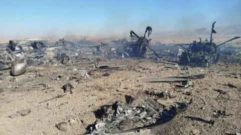 Zapaljene olupine američkih vojnih letjelica u pješčanoj pustinji Irana pod plavim nebom.